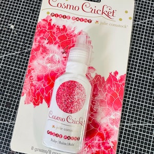 以下が含まれることがあります： ルビー色のCosmo Cricket Pixie Paintのボトル。ボトルは8グラムで、ピンクのグリッターが付いた白いラベルが付いています。