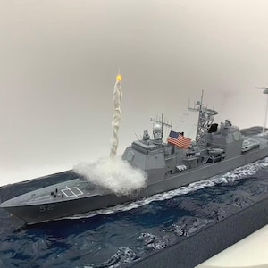 May include: A detailed model of a gray warship with the number "52" on the bow. The model features an American flag and a simulated missile launch with a plume of white smoke. The ship is set on a blue water base.