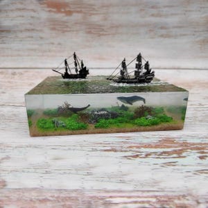 Puede incluir: Un diorama de resina que presenta una escena oceánica en miniatura. Dos veleros negros flotan en la superficie, mientras que una ballena y un barco hundido son visibles bajo el agua. Plantas verdes y arena completan el paisaje submarino.