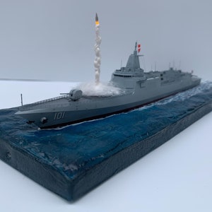 Puede incluir: Un modelo de barco gris con una estela de humo blanco detrás, disparando un misil. El barco está etiquetado con "101" y está sobre una base azul y gris.