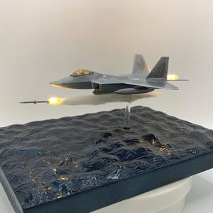 Può includere: Modello di aereo grigio con un missile in lancio, posto sopra una base d'acqua blu scuro strutturata. L'aereo ha un abitacolo giallo ed è montato su un supporto trasparente. La base quadrata poggia su una piattaforma rotante bianca.