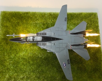 May include: A gray and white model airplane with the number 205 on the side and the number 502 on the tail. The airplane has a black cockpit and is sitting on a green surface. The airplane has two afterburners that are lit up.