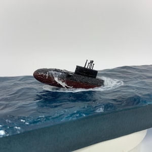May include: A black and red model submarine with a periscope, partially submerged in a blue resin ocean wave diorama.