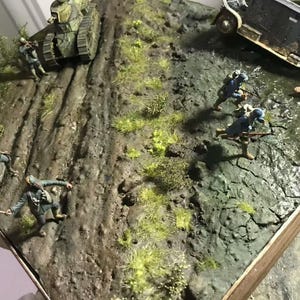 May include: A detailed diorama depicting a World War I battle scene. Includes a tank, armored car, soldiers in blue uniforms, and a muddy terrain with grass and water. The scene is set on a wooden base.
