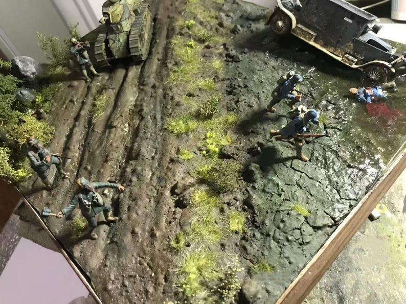 May include: A detailed diorama depicting a World War I battle scene. Includes a tank, armored car, soldiers in blue uniforms, and a muddy terrain with grass and water. The scene is set on a wooden base.