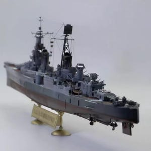 May include: A detailed model of a warship, predominantly gray with a red hull. The model features intricate details like turrets, antennas, and propellers. It is mounted on a gold-colored stand, showcasing a miniature replica of a naval vessel.