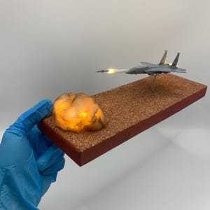 May include: A miniature model of a gray fighter jet with yellow accents, launching a missile. The jet is mounted on a brown rectangular base with a glowing rock formation. The base has a red border.