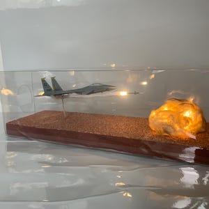 May include: A model airplane in flight, displayed inside a clear case. The model is gray and has a missile firing. A glowing, textured rock formation is on the right side of the display. The base is brown.