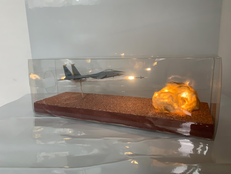 May include: A model airplane in flight, displayed inside a clear case. The model is gray and has a missile firing. A glowing, textured rock formation is on the right side of the display. The base is brown.