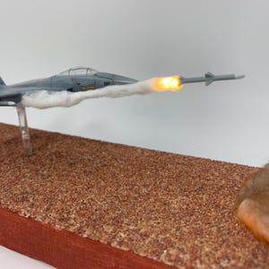 May include: A gray model jet plane with a missile firing, mounted on a brown base. The jet has the number 565 on the side. The missile has a bright orange glow at the end, with white smoke trailing behind the jet.