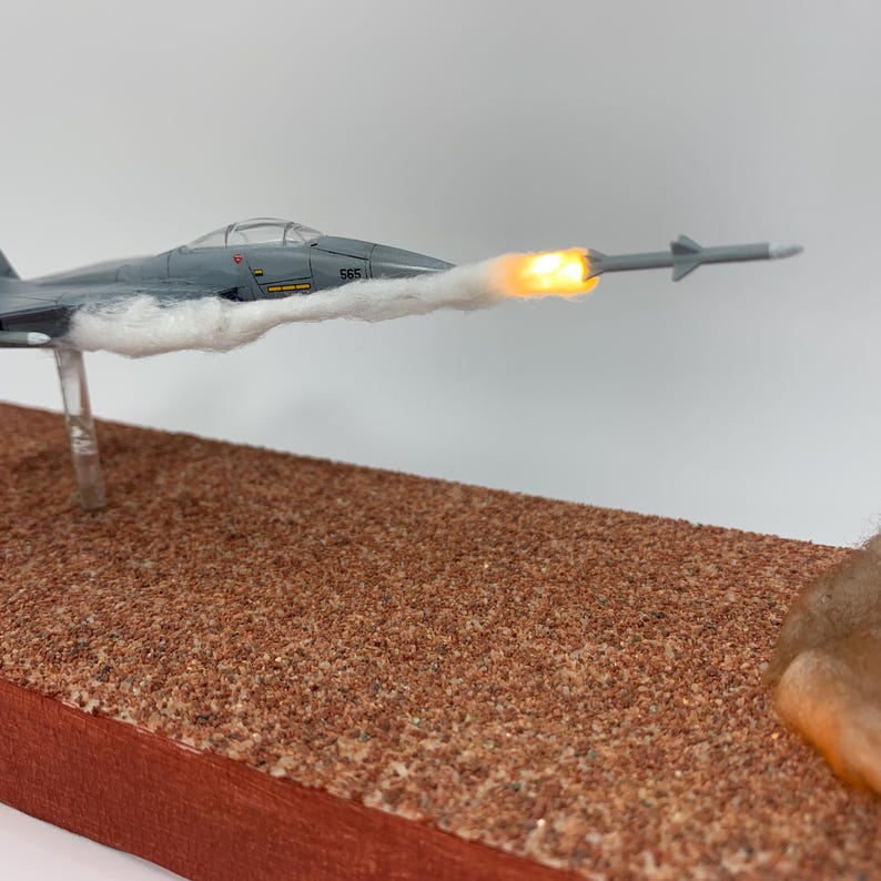 May include: A gray model jet plane with a missile firing, mounted on a brown base. The jet has the number 565 on the side. The missile has a bright orange glow at the end, with white smoke trailing behind the jet.