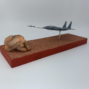 May include: A model airplane, painted in shades of gray, is mounted on a clear stand above a rectangular base. A missile is launched from the plane, leaving a trail of white smoke. A brown, textured object sits on the left side of the base.