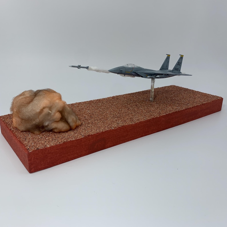 May include: A model airplane, painted in shades of gray, is mounted on a clear stand above a rectangular base. A missile is launched from the plane, leaving a trail of white smoke. A brown, textured object sits on the left side of the base.