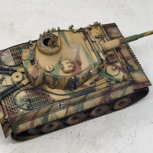 May include: A detailed model of a German Tiger I tank, painted in a camouflage pattern of green, brown, and tan. The tank has a raised gun barrel and is sitting on a white surface.