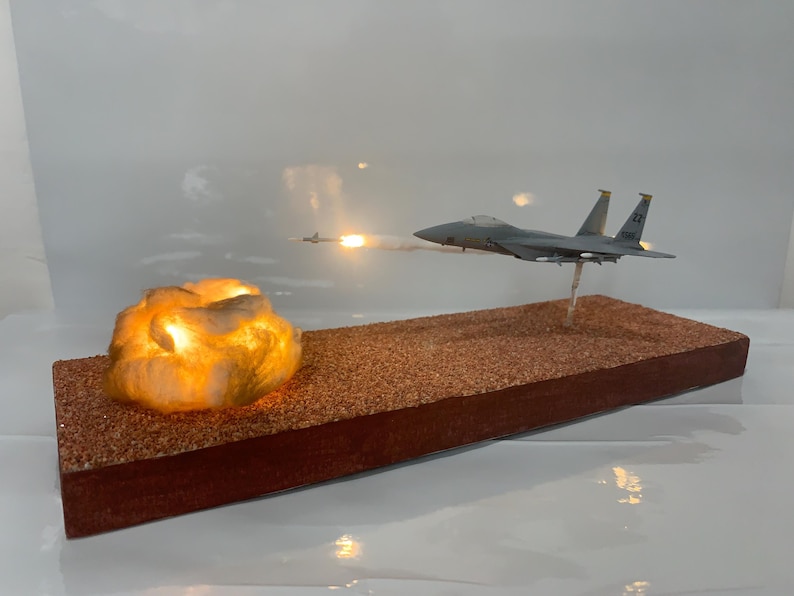 May include: A model airplane in flight, with a missile launch and a glowing cloud-like structure. The airplane is gray, and the base is brown with a reddish-brown border. The scene is set against a white background.