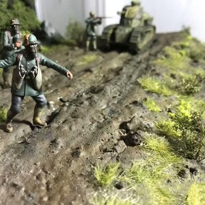 May include: A detailed diorama depicting a military scene with miniature soldiers, a tank, and terrain. The figures are dressed in green uniforms, and the tank is painted in camouflage. The terrain includes dirt, grass, and small bushes, creating a realistic battlefield.