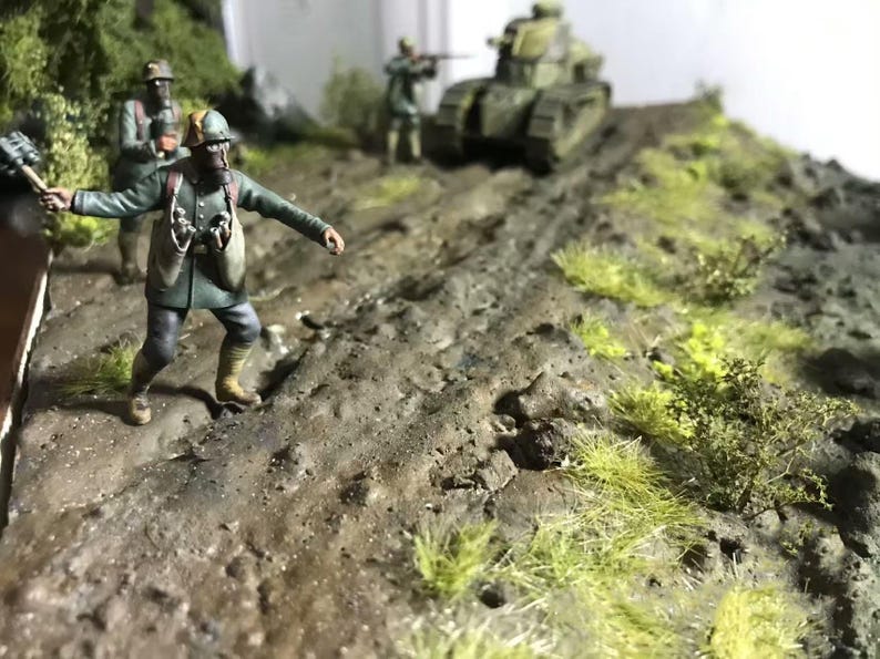 May include: A detailed diorama depicting a military scene with miniature soldiers, a tank, and terrain. The figures are dressed in green uniforms, and the tank is painted in camouflage. The terrain includes dirt, grass, and small bushes, creating a realistic battlefield.