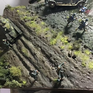 May include: A detailed diorama depicting a war scene with a tank, soldiers, and a vintage car on a textured, rocky terrain. The scene includes miniature figures in military uniforms, with green and brown tones dominating the landscape.