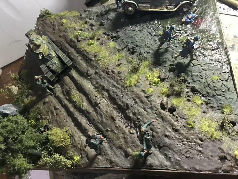 May include: A detailed diorama depicting a war scene with a tank, soldiers, and a vintage car on a textured, rocky terrain. The scene includes miniature figures in military uniforms, with green and brown tones dominating the landscape.