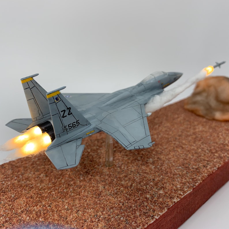May include: A detailed model of a gray F-15 fighter jet with yellow accents, emitting flames from its engines and launching a missile. The model is set on a brown, textured base, with a light-colored background.