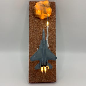 May include: A gray model jet plane with illuminated engines and a missile launch, mounted on a rectangular, textured brown surface. A glowing cloud-like element is at the top. The piece is lit with warm-toned lights.