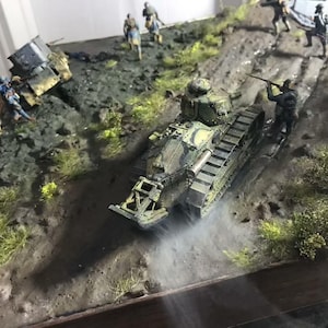 May include: A detailed diorama depicting a military scene with a camouflaged tank as the central focus. Soldiers are positioned around the tank and on a dirt path. The scene includes miniature trees and foliage, creating a realistic battlefield setting.