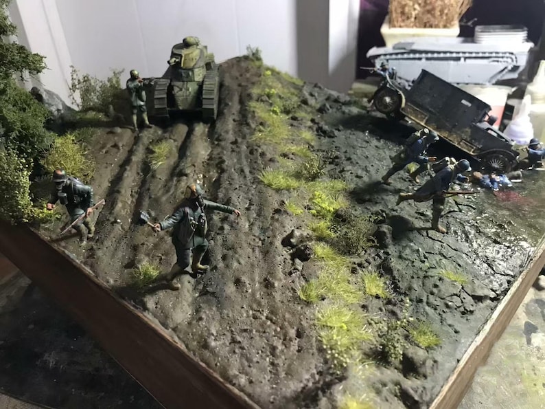 May include: A detailed diorama depicting a World War I battle scene. A tank ascends a muddy hill, while soldiers and a vehicle are positioned on the terrain. The scene is set with miniature figures and foliage, creating a realistic war environment.
