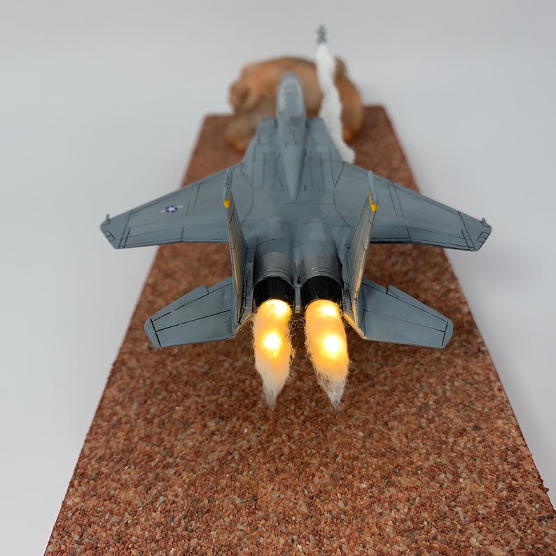 May include: A gray model airplane with illuminated engine exhaust, positioned on a textured brown surface. The miniature jet has detailed wings and a tail, with a plume of white smoke rising from the back. The model is a detailed replica.
