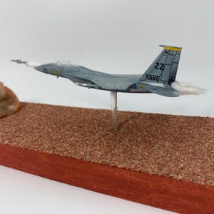 May include: A gray model airplane with the markings "ZZ" and "565" on the tail, mounted on a clear stand. The model is positioned above a brown, textured surface, with a red wooden base. White vapor trails extend from the plane's rear.