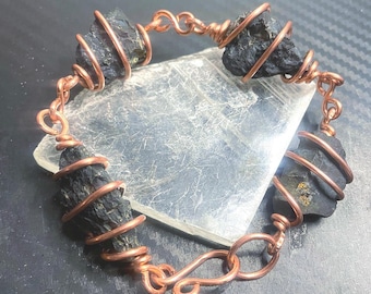 May include: A bracelet featuring three raw, dark black stones wrapped in copper wire. The stones are connected by copper wire links, with a hook-and-eye clasp mechanism. The copper wire is coiled around each stone, creating a series of loops.