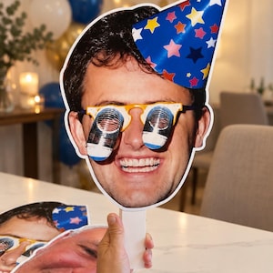 May include: A large, die-cut cardboard cutout of a smiling face wearing a blue party hat with colourful stars and yellow-rimmed shutter glasses. The cutout is attached to a white handle, and a second, partial face cutout is visible at the bottom.
