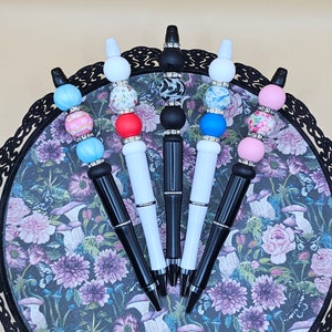 May include: Six black pens with white and black decorative beads. The beads have various patterns including floral, geometric, and abstract designs. The pens are arranged in a fan shape on a black and white floral tray.