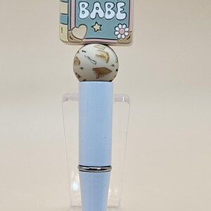 May include: A light blue pen with a book charm. The charm is decorated with a pink, yellow, and green book with the text "BOOK BABE" in white. The pen is on a clear acrylic stand.