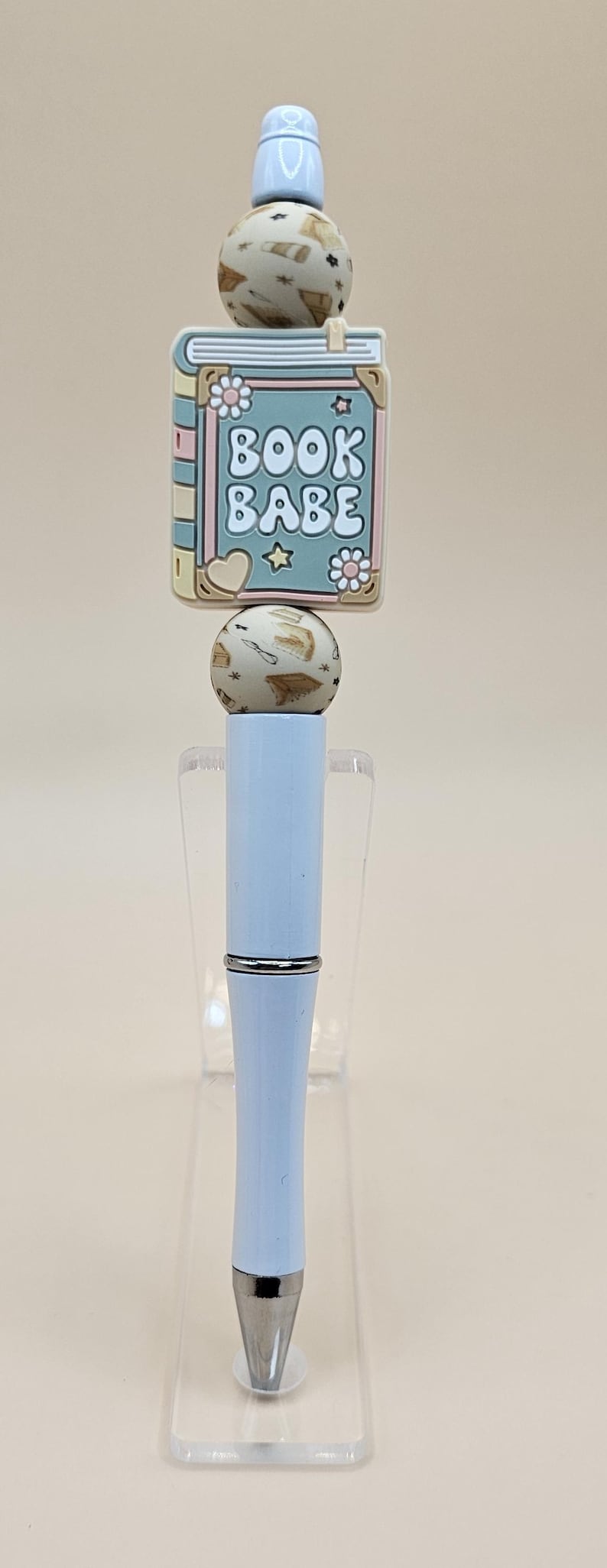 May include: A light blue pen with a book charm. The charm is decorated with a pink, yellow, and green book with the text "BOOK BABE" in white. The pen is on a clear acrylic stand.