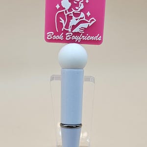 May include: A white pen with a pink charm that says "Crying Over Book Boyfriends". The charm features a white illustration of a person reading a book.