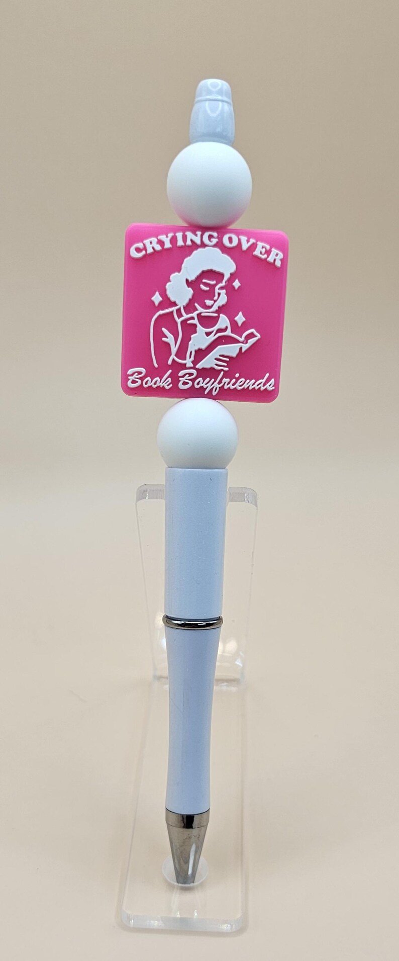 May include: A white pen with a pink charm that says "Crying Over Book Boyfriends". The charm features a white illustration of a person reading a book.