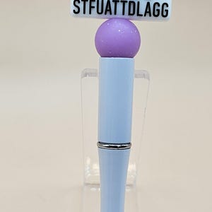 May include: A purple and white pen with a charm that says "Buy Me Books and Tell Me to STFUATTDLAGG".