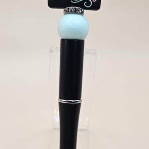 May include: Black pen with a light blue and black beaded top. The bead has a black chalkboard with the text "CRAZY BOOK Lady" in rainbow colors.