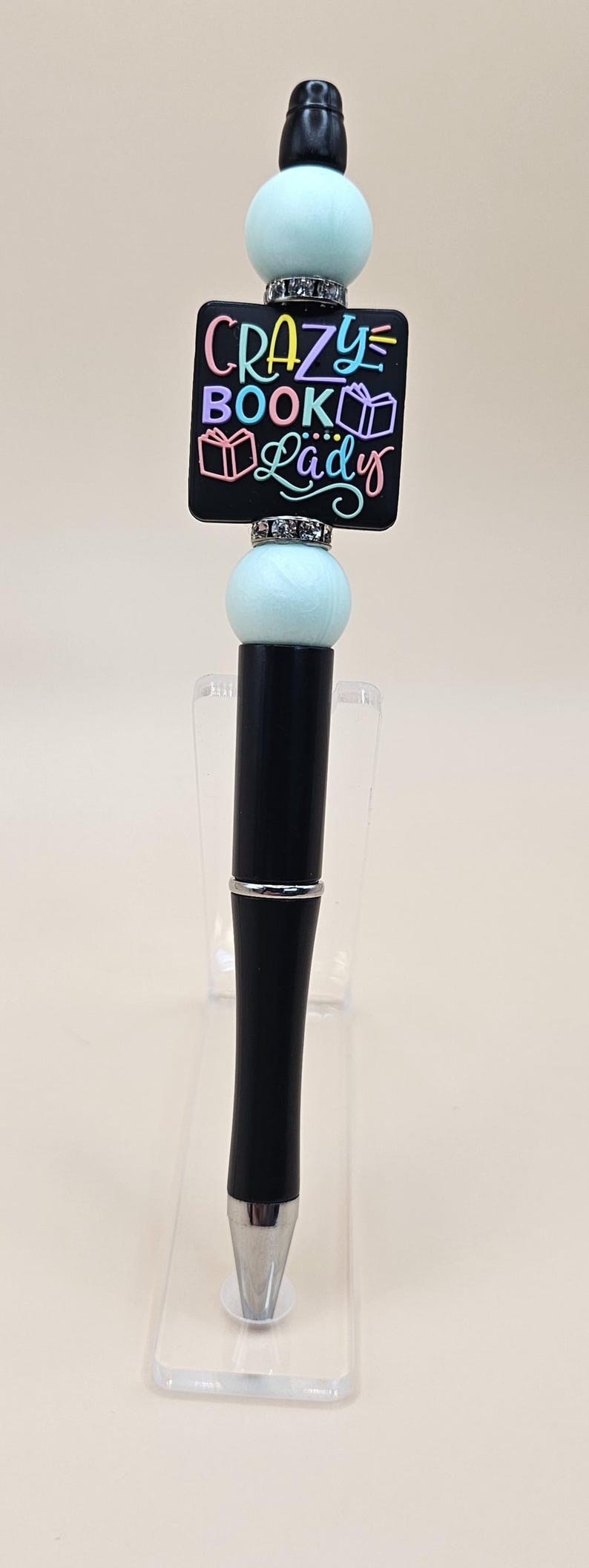 May include: Black pen with a light blue and black beaded top. The bead has a black chalkboard with the text "CRAZY BOOK Lady" in rainbow colors.