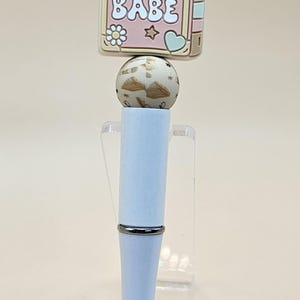 May include: A white pen with a silver band and a pink and beige book charm with the text "BOOK BABE".
