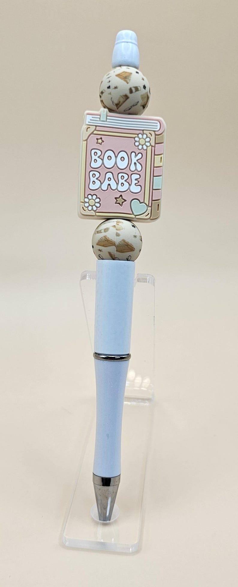 May include: A white pen with a silver band and a pink and beige book charm with the text "BOOK BABE".