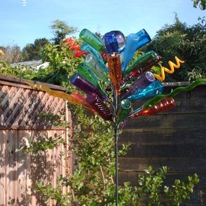 May include: A colorful glass bottle wind chime with a metal frame and a variety of colored glass bottles. The wind chime is hanging from a metal pole and is surrounded by green foliage.