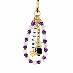 May include: A gold-toned keychain featuring a teardrop-shaped loop of alternating purple and white beads with small iridescent star spacers. Dangling from the center are a white bead strand spelling TIGERS, a yellow heart charm, and a football charm.
