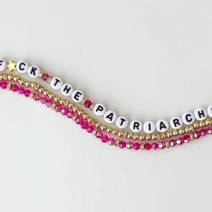 Puede incluir: Tres pulseras de cuentas en capas con la frase "F*CK THE PATRIARCHY" escrita con cuentas de letras blancas. Las pulseras presentan cuentas doradas y rosas, con un dije de estrella dorada.