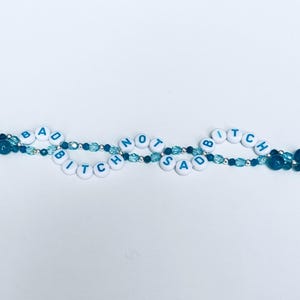 May include: A beaded necklace with the words "BAD BITCH NOT SAD" spelled out in white letter beads. The necklace features blue and silver beads, creating a two-strand design against a white background.