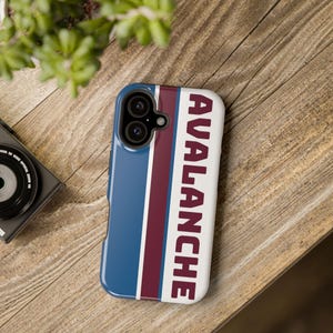 May include: A blue, white, and burgundy phone case with the word "AVALANCHE" in burgundy vertical text. The case has a blue stripe on the left side and burgundy and white stripes on the right. The case is on a wooden surface.
