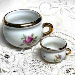 May include: Two white porcelain cups with gold rims and handles. The larger cup features a pink rose illustration, while the smaller cup has a floral design and the word "Cuit". Both cups are on a white lace surface.