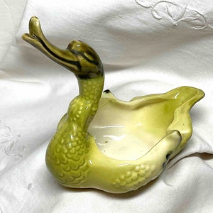 May include: A green ceramic duck planter with a textured surface and a brown head. The duck's head is bent back and the planter is shaped like a duck's body.