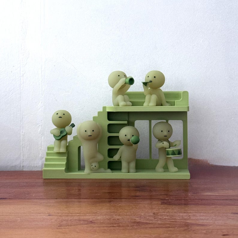 May include: A quirky, pale green miniature stage set featuring six small figures. Some figures play instruments, including a guitar, trumpet, and drums. The figures are positioned on a two-tiered structure with stairs and a window.