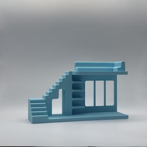 May include: A light blue architectural model featuring stairs, shelves, and a window-like aperture. The model has a flat base and a rectangular top. The design is minimalist and geometric, with clean lines and a solid colour.